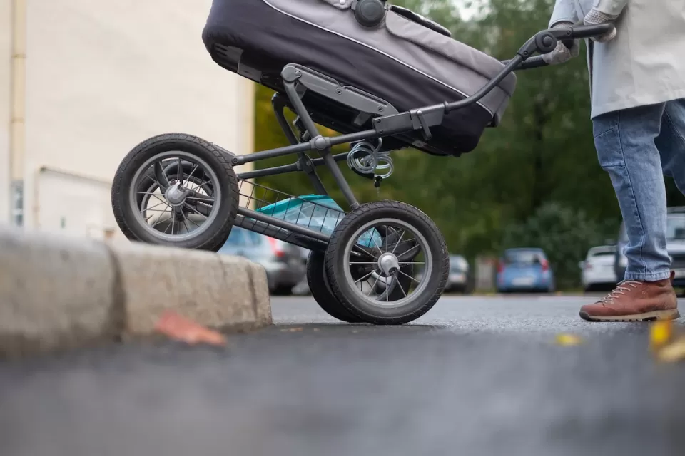 Close-up of bottom half of stroller being pushed onto sidewalk with no curb cut by person in jeans and brown shoes.